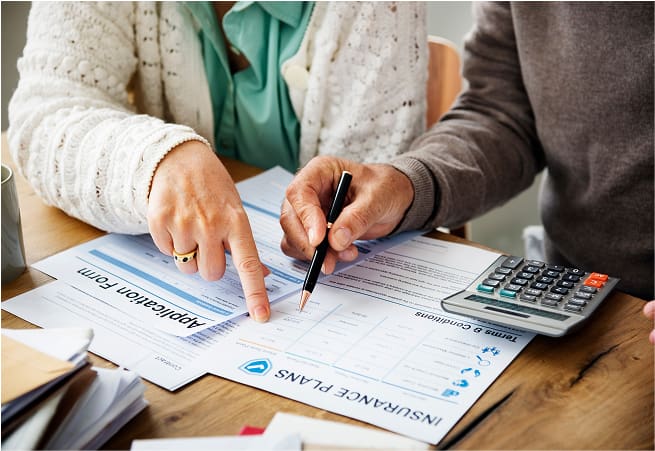 Two people are reviewing an insurance plan document on a wooden table, with one person pointing at a section and the other holding a pen. There is also a calculator and a stack of papers nearby.