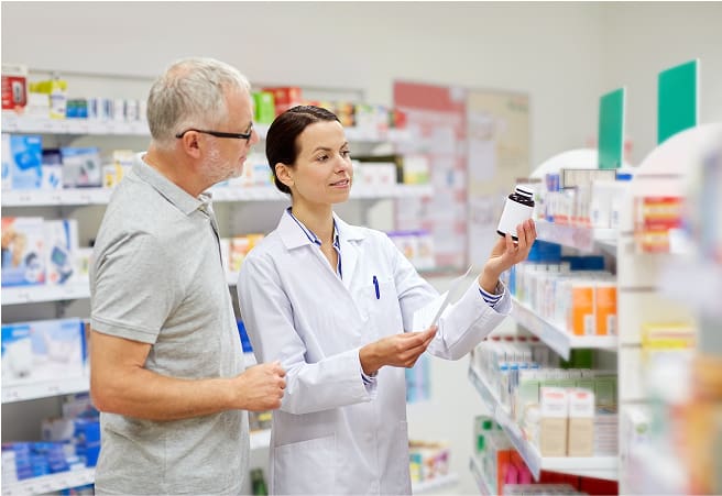 A young female pharmacist in a white lab coat shows a bottle of medication to an older male customer in a pharmacy aisle filled with various medicine boxes and bottles.
