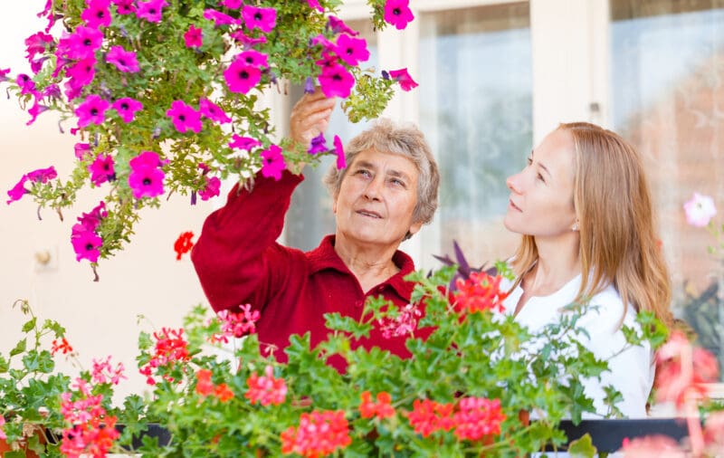 An elderly woman and a younger woman are in a garden surrounded by colorful pink and red flowers. The elderly woman is reaching up to touch a hanging pink flower, while the younger woman looks on attentively. The scene appears peaceful and nurturing.