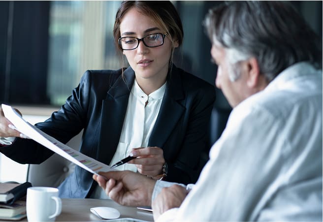 A young woman in business attire and glasses reviews documents with an older man at a table in an office setting.