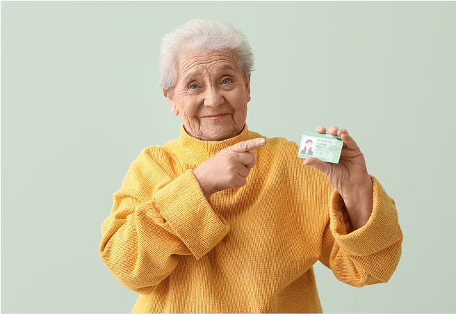 An elderly woman with short white hair, wearing a yellow sweater, smiling and pointing to a small card she is holding in her right hand.