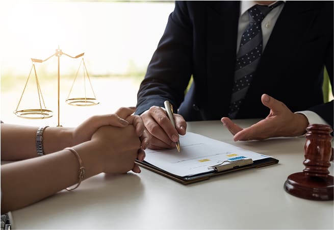 A person in a suit and tie is sitting at a desk, holding a pen and writing on a document, while another person's hands are clasped on the desk. A gavel and a set of scales are visible on the desk, indicating a legal or counseling setting.