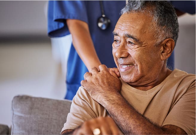 An elderly man sits on a couch, smiling, as a healthcare worker in blue scrubs adjusts his shoulder. The healthcare worker's face is not visible.